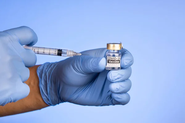 Close-up of hand with surgical glove holding covid-19 vaccine vial on blue background — Stock Photo, Image