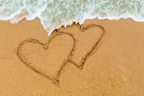 Двойное сердце, нарисованное на песчаном пляже с приближающейся волной
