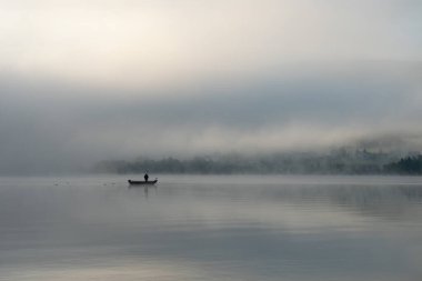 İsviçre 'nin Lac de Joux şehrinde sabahın erken saatlerinde: Teknede yalnız bir balıkçı, sis hala yükselen güneşi saklıyor