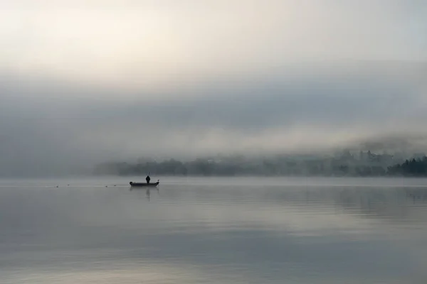 İsviçre 'nin Lac de Joux şehrinde sabahın erken saatlerinde: Teknede yalnız bir balıkçı, sis hala yükselen güneşi saklıyor