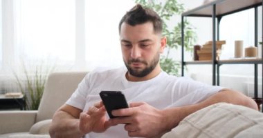 Akıllı telefon kullanan yakışıklı yetişkin adam evdeki kanepede oturuyor.