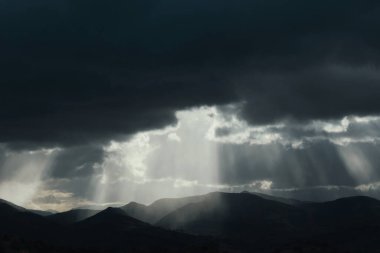 Gün batımında dramatik gökyüzü, bulutlarda güneş ışığı ve ufukta dağlar.