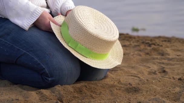 Woman sits in sandy lake side with straw hat — Stock Video