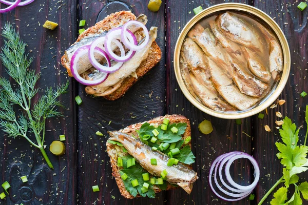 Зерновой хлеб с сардинами и зеленью рядом с банком шпроты на черном фоне с укропом и петрушкой — стоковое фото