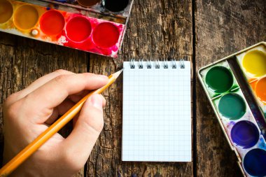 Hand of the artist with a brush for drawing on a background of old table, watercolors and notepad selective focus