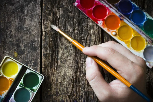 Hand of the artist with a brush for drawing on a background of the old table and watercolors selective focus