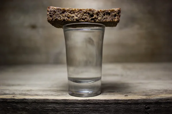 Un vaso de vodka y una rebanada de pan de centeno en una mesa de madera . — Foto de Stock