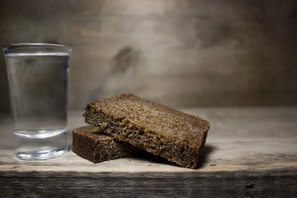 Un vaso de vodka y una rebanada de pan de centeno en una mesa de madera . — Foto de Stock