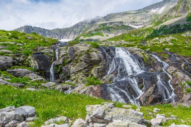 Dağ vadisinde, çimenli kıyıların arasında hızlı bir dere var. Yeşil çayırda küçük şelale.
