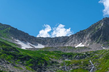 Mavi gökyüzünün altında kar ve taş yerleşimli kaya sırtı. Dağ vadisine yaz gezisi. Atmosferik alp manzarası yeşil çimenli taşlı çayır.