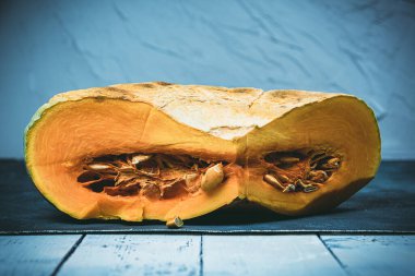 Büyük bir parça balkabağı ikiye bölünmüş. Mavi arka planda ahşap masada yemek pişirmek için turuncu sebze. Balkabağının içinde sulu hamur ve tohumlar.