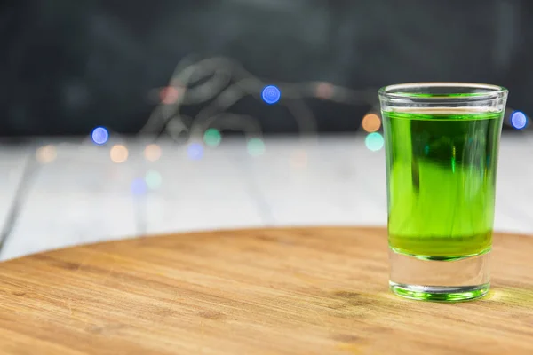 Green drink on wooden bar. Shot glass of clear absinthe. Festive atmosphere, a colorful garland in blur. Drinking alcohol alone. Copy space for text