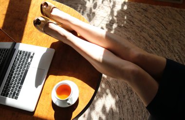 Bacakları masanın üzerinde olan olgun bir kadın - bitki çayı ve dizüstü bilgisayar. 