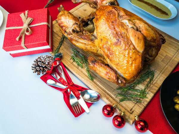 Целая жареная индейка и розмарин на деревянной разделочной доске с праздничным оформлением и столовые приборы вокруг него, пюре картофельное и брюссельская капуста на спине на белой скатерти и красный стол бегун
