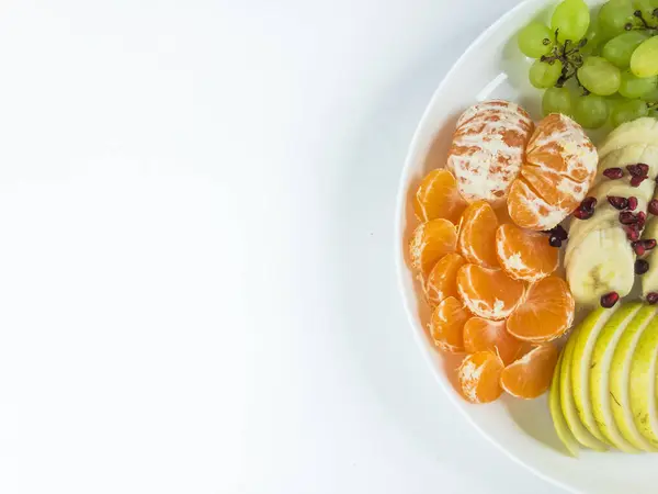 İzole edilmiş beyaz bir zemin üzerinde beyaz bir plaka içinde muz, üzüm, armut, mandalina ve nar karıştırılmış taze meyveler.