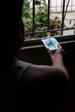 İspanyol bir adam pencerenin önünde oturuyor. Elinde bir n95 cerrahi maske var. Arkadan görülüyor.