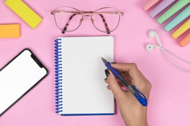 Pembe arka planda boş bir defter, boş ekranlı bir cep telefonu, renkli post-it notları, kulaklıklar, renkli kalemler, gözlükler ve bir şeyler yazacak bir el.