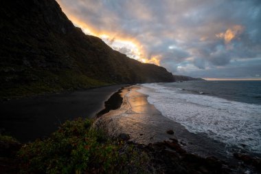 Renkli gün batımı, bulutlu bir gökyüzü ile kara kumlu bir sahilde deniz suyuna yansıyor.