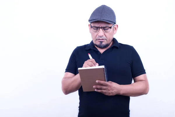 Man on a white background in a cap, glasses, takes notes with a pen and a notebook.