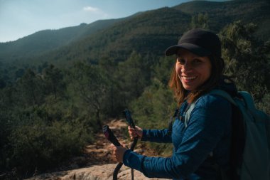 Bir yürüyüşçü kep takar ve baston taşır. Etrafı doğa ile çevriliyken gülümser..