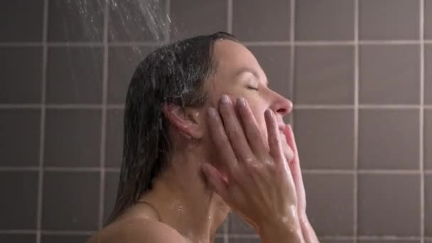 Atractiv bruneta femeie de vârstă mijlocie ia un duș. Femeia dezbrăcată îşi spală părul. Țiglă gri pe pereți. Îngrijirea pielii, tratamente de rutină la domiciliu . — Videoclip de stoc