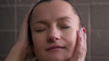 Çekici esmer, orta yaşlı kadın duş alıyor. Çıplak kadın saçlarını yıkıyor. Duvarlarda gri fayans var. Cilt bakımı, rutin ev tedavileri.