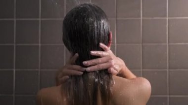 Çekici esmer, orta yaşlı kadın duş alıyor. Çıplak kadın saçlarını yıkıyor. Duvarlarda gri fayans var. Cilt bakımı, rutin ev tedavileri.