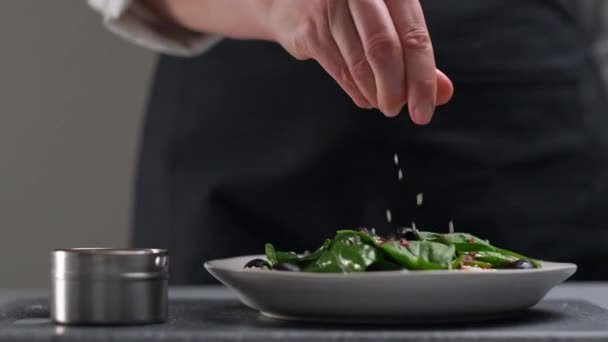 En kvinnlig kock i vit uniform och ett svart förkläde i restaurangköket. Kocken strör salladen med sesamfrön. — Stockvideo