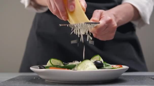 Una chef con uniforme blanco y un delantal negro en la cocina del restaurante. Cocinar una ensalada. El cocinero frota el queso parmesano sobre un pequeño rallador. — Vídeos de Stock