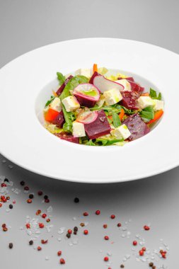 Turplu sebze salatası, yumuşak peynir, pancar kökü ve gri arka planda beyaz bir tabakta yeşillikler. Lokantada atıştırmalıklar.