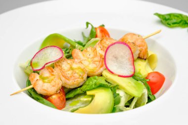 Şişte ızgara karides ve gri arka planda beyaz tabakta sebzeler..