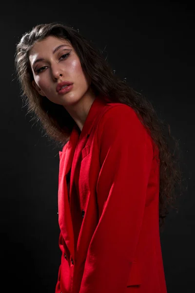 Een jonge mooie oosterse vrouw met lang zwart haar in een rood jasje tegen een zwarte achtergrond. Het modemodel poseert in de studio. — Stockfoto