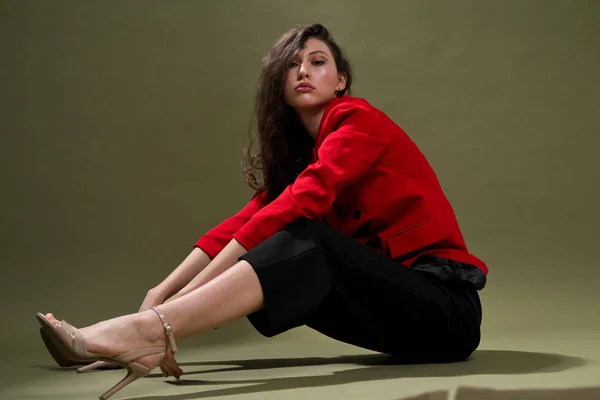 A sensual young woman in a red jacket, culottes and shoes with wet long black hair poses against a green background in the studio.