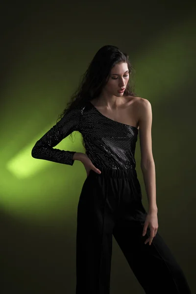 A beautiful young tall girl with long black hair poses in a shiny blouse and black culottes on a green background. Buty and fashion photo shoot. — Stock Photo, Image