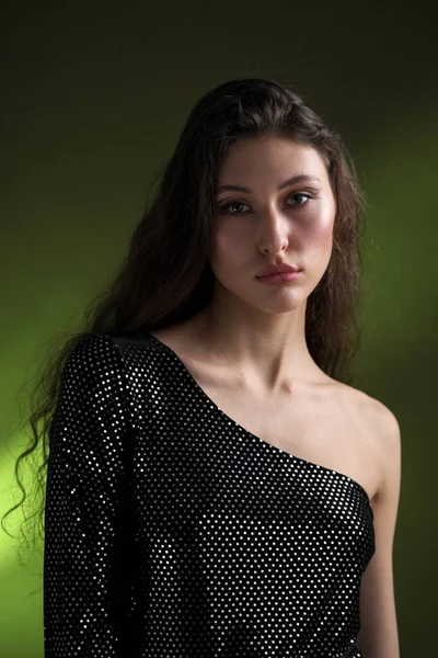 A beautiful young tall girl with long black hair poses in a shiny blouse and black culottes on a green background. Buty and fashion photo shoot. — Stock Photo, Image