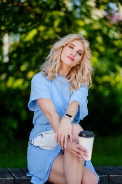 Mavi kısa elbiseli ve beyaz kemer çantalı güzel sarışın kız yeşil bir parkta yürüyor. Bankta oturur ve kahve içer..