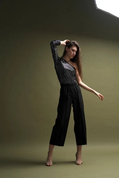 Una hermosa joven alta con el pelo largo y negro posa en una blusa brillante y culottes negros sobre un fondo verde. Buty y sesión de fotos de moda. — Foto de Stock