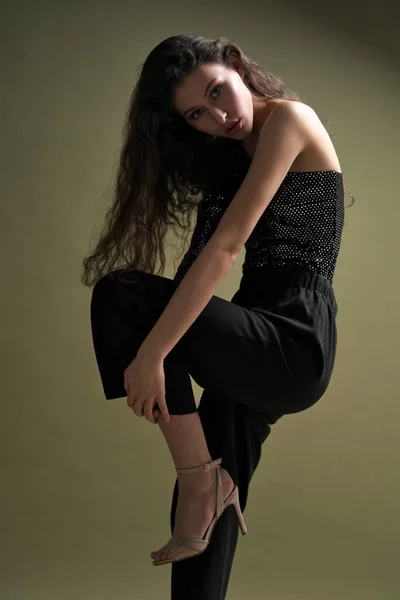 A beautiful young tall girl with long black hair poses in a shiny blouse and black culottes on a green background. Buty and fashion photo shoot. — Stock Photo, Image