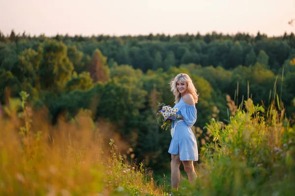 Ritratto di una giovane bella ragazza bionda vestita di blu. Capelli sciolti, labbra sensuali e grandi occhi azzurri. Una donna tiene un mazzo di margherite fiori di campo . — Foto Stock