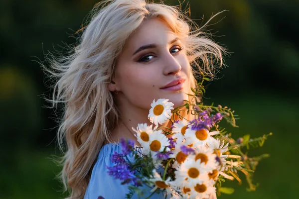 Portret młodej pięknej blondynki w niebieskiej sukience. Luźne włosy, zmysłowe usta i duże niebieskie oczy. Kobieta trzyma bukiet stokrotek dzikie kwiaty . — Zdjęcie stockowe