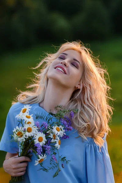Porträtt av en ung vacker blond flicka i en blå klänning. Löst hår, sensuella läppar och stora blå ögon. En kvinna håller en bukett prästkragar vildblommor . — Stockfoto