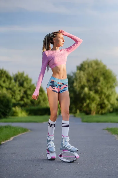 Eine junge, schlanke, schöne Frau mit Dreadlocks in Sportkleidung trainiert draußen im Park. Kangoo springt beim Training. — Stockfoto