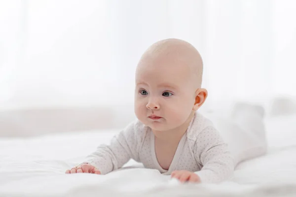 Portrait d'un beau bébé mignon aux yeux gris en pyjama blanc sur le lit contre la fenêtre. — Photo