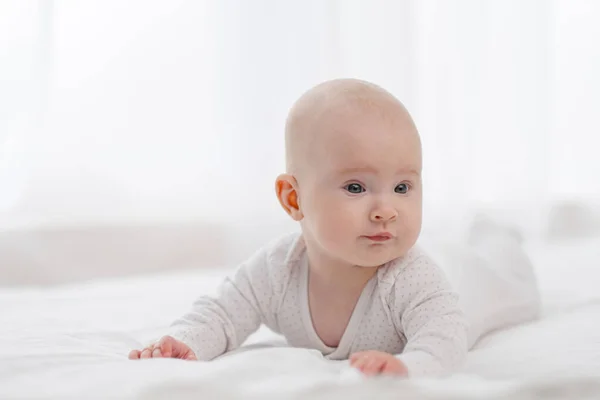 Portrait of a cute beautiful baby with gray eyes in white pajamas on the bed against the window. — Stock Photo, Image