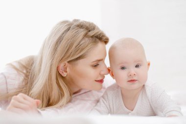 Genç, güzel, beyaz, sarışın anne bebeğiyle birlikte yatakta yatıyor. Beyaz ışıklı ev içi.
