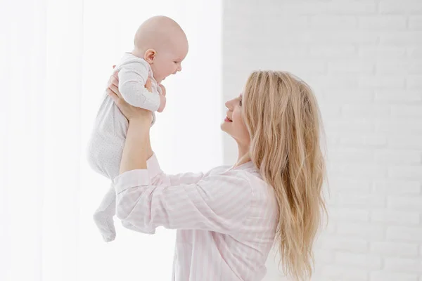 Una giovane madre caucasica in abiti da casa tiene in braccio un neonato con una tuta bianca. Sorrisi e abbracci il bambino. — Foto Stock