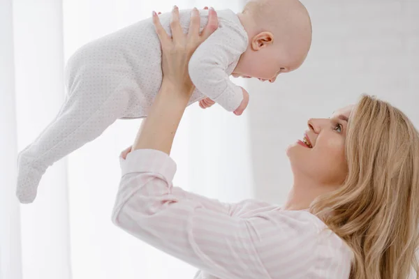 Una giovane madre caucasica in abiti da casa tiene in braccio un neonato con una tuta bianca. Sorrisi e abbracci il bambino. — Foto Stock