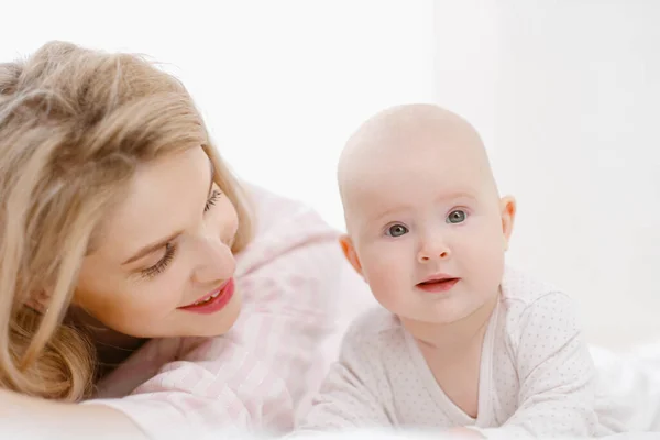 Giovane bella mamma bionda caucasica si trova sul letto con la sua bambina. Luce bianca interno della casa. — Foto Stock