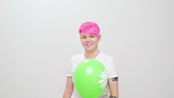 Young beautiful girl with a short haircut and bright hair, painted pink. A model poses on a white background, holds a green balloon in hands. – Stock-video