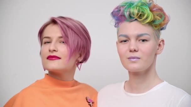 Deux jeunes filles aux cheveux teints brillants et aux coupes de cheveux courtes. Coiffeur coloriste et mannequin pose sur un fond blanc. Couple homosexuel lesbienne. — Video